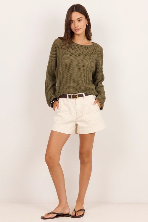 A woman modeling a long-sleeve olive green knit top, cream shorts, a thin brown belt, and black flip-flops against a plain off-white studio background.