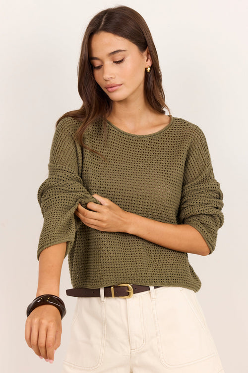 A woman wearing a textured olive green knit long-sleeve top and cream shorts, standing against a plain off-white background.