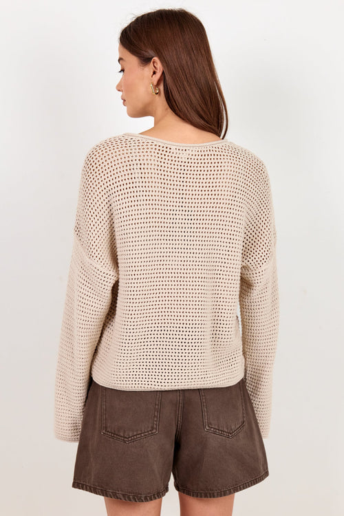 A woman shown from the back wearing a cream-colored, open-knit long-sleeve sweater and brown shorts against a plain off-white background.