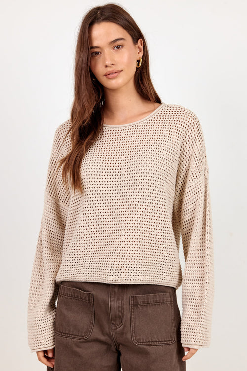A woman modeling a beige open-knit long-sleeve sweater and dark brown shorts against a plain light background.