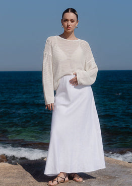 Woman in a white sweater and skirt standing on a rocky shore with ocean and clear blue sky in the background