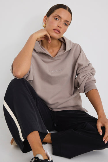 Woman wearing a relaxed-fit taupe hoodie with subtle logo embroidery, styled with black wide-leg pants featuring a white side stripe.