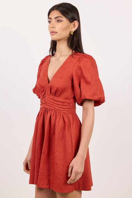 A woman modeling a terracotta orange short puff-sleeve dress with a gathered waist and deep V-neckline, photographed against a plain off-white studio background.