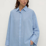 A woman wearing a light blue long-sleeve button-up shirt with subtle vertical stripes, paired with light-wash blue jeans, standing against a plain off-white background.