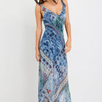 Woman wearing a blue maxi dress with a paisley scarf print, ruffle shoulder straps, and flowing bias-cut silhouette.