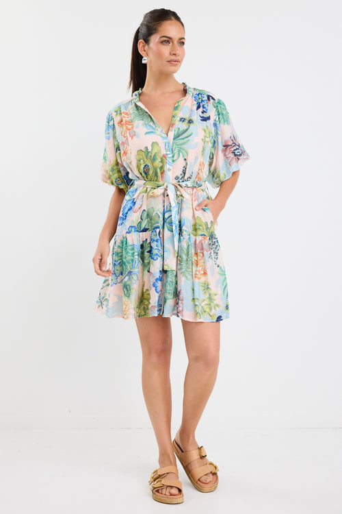 Woman wearing a colourful floral mini dress with a waist tie, short sleeves, and a tiered skirt.







