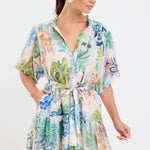 Woman wearing a colourful floral mini dress with a waist tie, short sleeves, and a tiered skirt.







