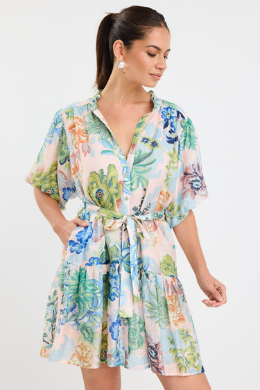 Woman wearing a colourful floral mini dress with a waist tie, short sleeves, and a tiered skirt.







