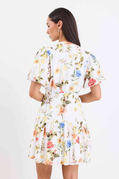 Woman wearing a cream floral mini dress with a tie belt, button front, short puff sleeves, and a drop waist design.