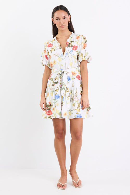 Woman wearing a cream floral mini dress with a tie belt, button front, short puff sleeves, and a drop waist design.