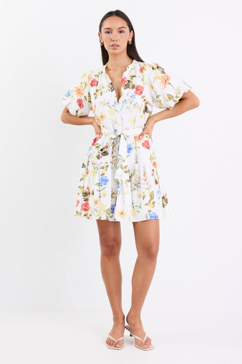 Woman wearing a cream floral mini dress with a tie belt, button front, short puff sleeves, and a drop waist design.
