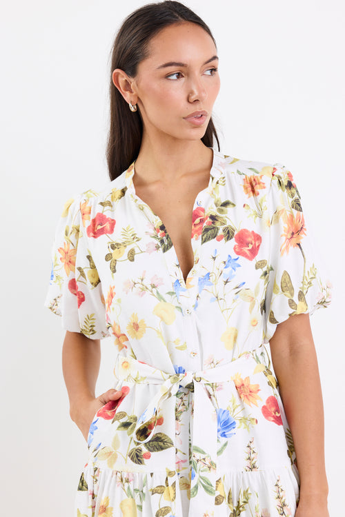 Woman wearing a cream floral mini dress with a tie belt, button front, short puff sleeves, and a drop waist design.