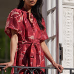 A woman stands on a balcony wearing a red patterned short-sleeve dress with a tie waist. The dress has a pinkish-red base with lighter abstract floral or marbled patterns.