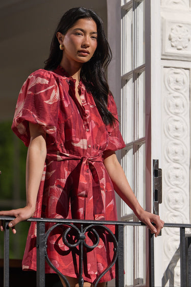 A woman stands on a balcony wearing a red patterned short-sleeve dress with a tie waist. The dress has a pinkish-red base with lighter abstract floral or marbled patterns.
