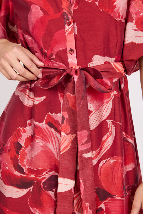 A close-up of a red floral dress with a matching fabric tie belt, featuring a large watercolor-style flower print in shades of red, pink, and white.