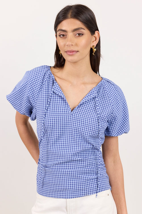 A woman modeling a blue and white gingham short-sleeve blouse with a split V-neckline and ruffled shoulders, paired with white pants, against a plain off-white background.