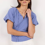 A woman modeling a blue and white gingham wrap-style blouse with short puff sleeves and a split neckline, paired with white pants, against a plain light background.