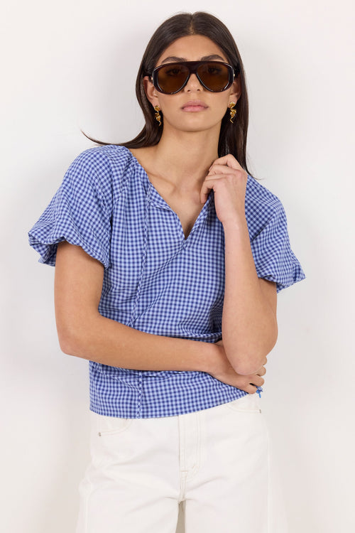A woman modeling a blue and white gingham wrap-style blouse with short puff sleeves and a split neckline, paired with white pants, against a plain light background.