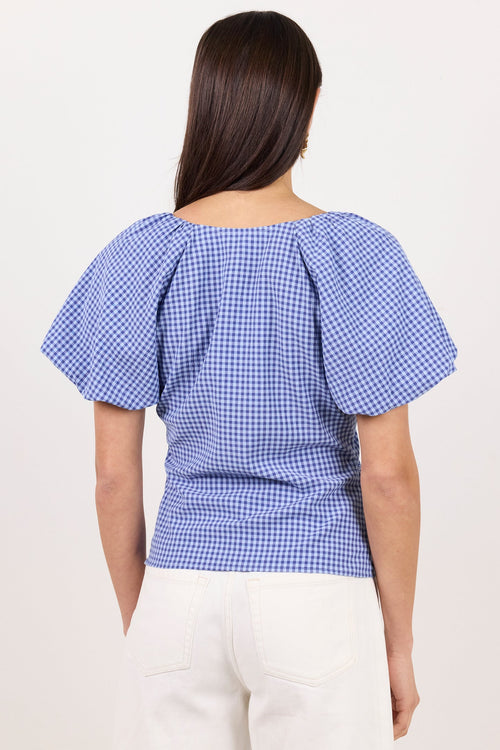 A woman viewed from the back wearing a blue and white gingham short-sleeve top with puff sleeves and white pants, set against a plain off-white studio background.
