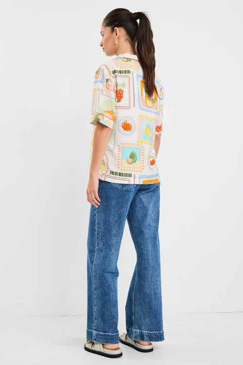 Woman wearing a colourful short sleeve blouse with postcard-style fruit prints and a button front, styled with blue jeans.