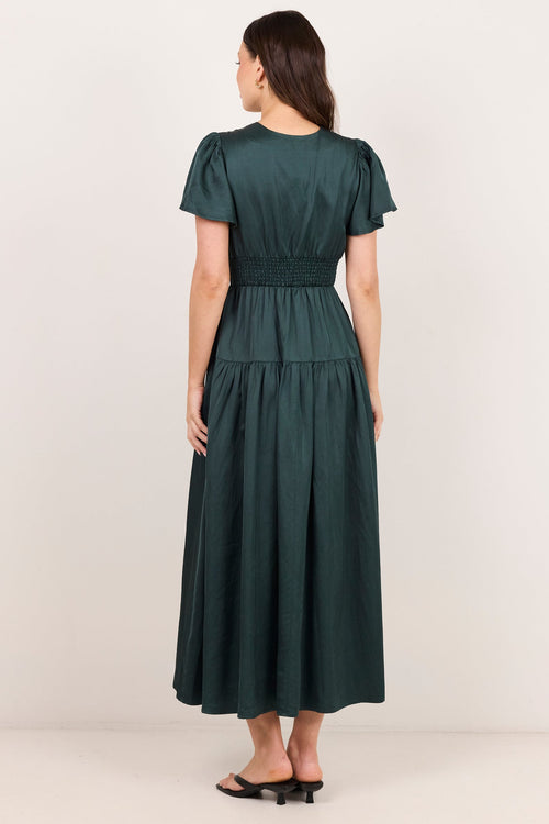 A woman viewed from the back wearing a dark green midi dress with short puff sleeves, a smocked waist, and a tiered skirt, paired with black flat sandals, against a plain off-white studio background.