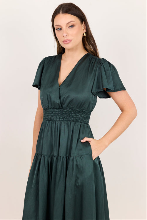 A woman modeling a dark green midi dress with short flutter sleeves, a smocked waist, and a tiered skirt against a plain light background.