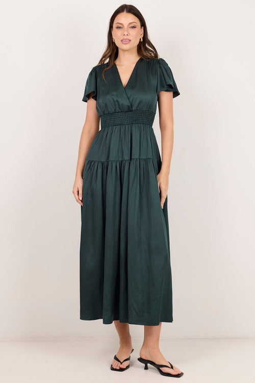 A woman modeling a dark green midi dress with short flutter sleeves, a V-neckline, and a peplum waist, paired with black flip-flop sandals against a plain off-white studio background.