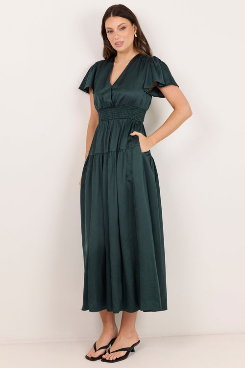 A woman modeling a dark green midi dress with short flutter sleeves, a V-neckline, and side pockets, standing against a plain off-white studio background.