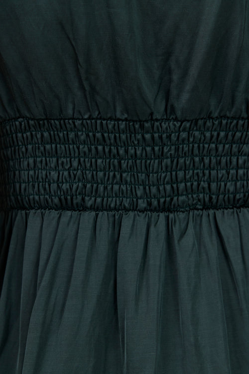 Close-up of a dark forest green smocked dress fabric, showing the gathered elastic waistband and draped skirt texture.