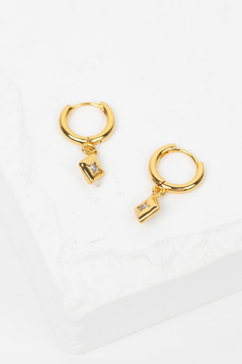 A pair of gold huggie hoop earrings with small rectangular clear gemstone charms, displayed on a white textured stone block.