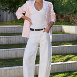 A woman stands on outdoor stone steps wearing a light pink blazer, white tank top, white wide-leg pants, a black belt, and black sandals, with green foliage in the background.