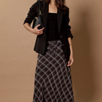 A woman models a black blazer, black top, and a black and white plaid maxi skirt against a solid beige studio backdrop.