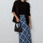 A woman stands against a white brick wall wearing a black blazer, black top, a blue plaid maxi skirt, black flats, and carrying a black handbag.