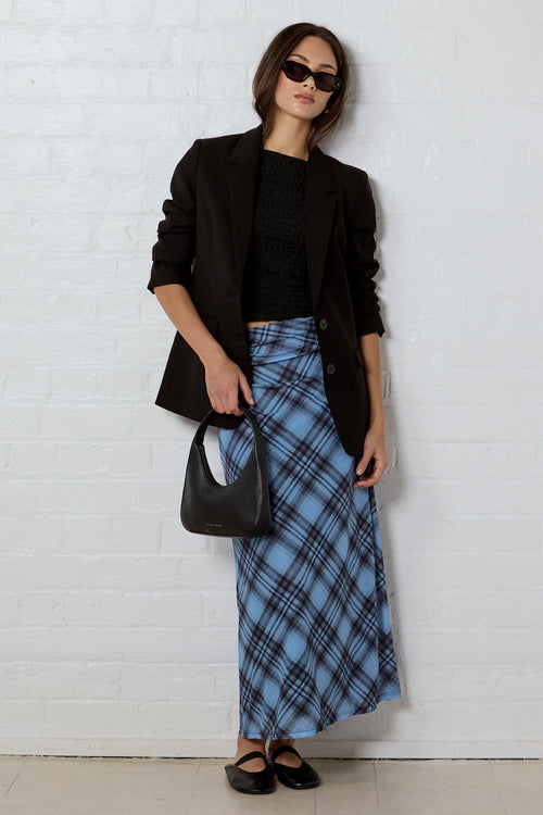 A woman stands against a white brick wall wearing a black blazer, black top, a blue plaid maxi skirt, black flats, and carrying a black handbag.