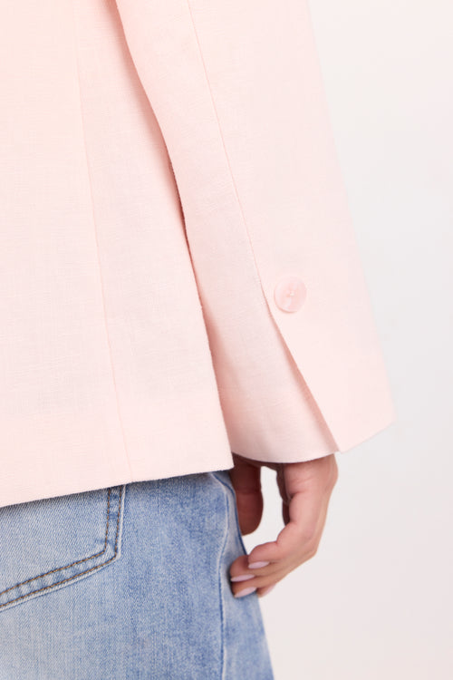 Close-up of a person wearing a light pink blazer with a single visible button and blue jeans against a plain light background.
