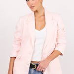 A woman modeling a light pink blazer over a white tank top and blue jeans, standing against a plain white studio background.