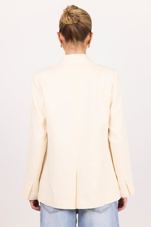 A woman viewed from behind wearing a light cream-colored blazer and light blue jeans against a plain white studio background.