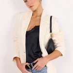 A woman posing in a cream-colored blazer over a black lace-trimmed camisole top and light-wash blue jeans, carrying a black shoulder bag with gold hardware against a plain white background.