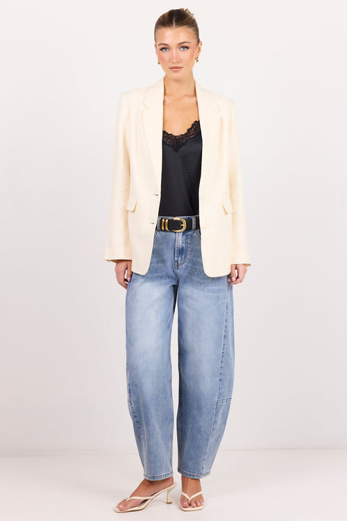 A woman stands against a plain white studio background wearing a cream-colored blazer, a black lace-trimmed top, wide-leg light-wash blue jeans, and beige heeled sandals.