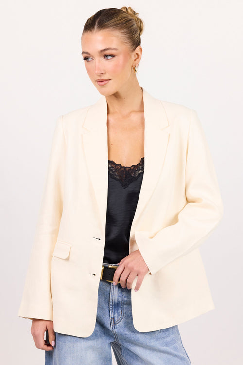 A woman modeling a cream-colored blazer over a black lace-trimmed top and light-wash blue jeans, standing against a plain white studio background.