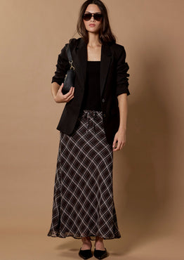 A woman stands against a plain beige studio backdrop wearing a black blazer, black top, a long black-and-white plaid skirt, black pointed shoes, and sunglasses, while holding a small black handbag.
