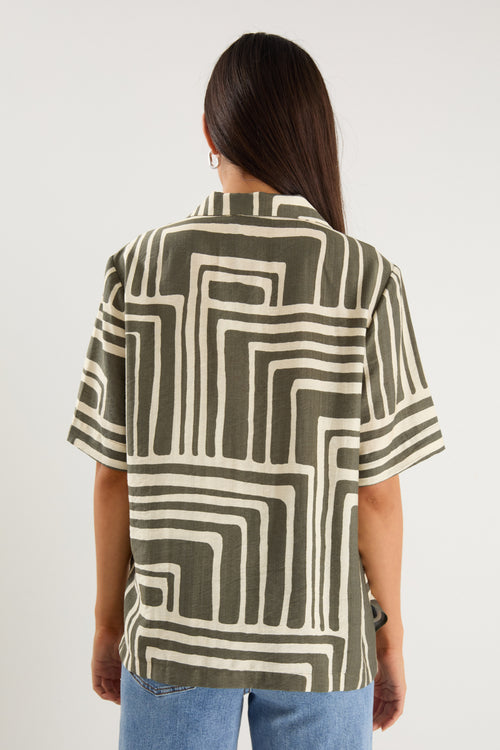 Woman wearing a short-sleeve shirt with an abstract olive and cream geometric print and button front, styled with mid-wash denim jeans.