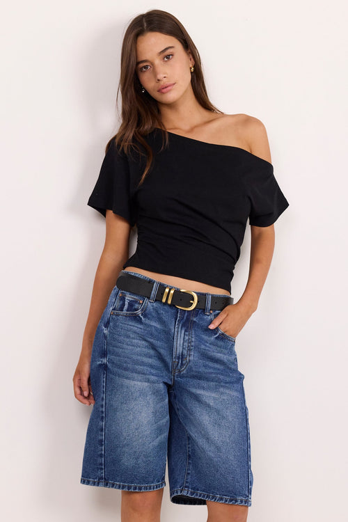A woman modeling a black off-the-shoulder short-sleeve crop top and medium-wash blue denim shorts with a black belt, standing against a plain white studio background.