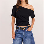 A woman modeling a black off-the-shoulder short-sleeve crop top paired with blue denim shorts and a black belt with a gold buckle, against a plain light beige studio background.