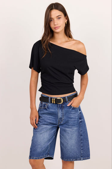 A woman modeling a black off-the-shoulder short-sleeve crop top paired with blue denim shorts and a black belt with a gold buckle, against a plain light beige studio background.