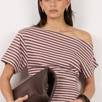 A woman stands against a plain off-white studio wall wearing a striped off-the-shoulder top, dark brown cropped trousers, and black heeled sandals, while holding a dark brown clutch.