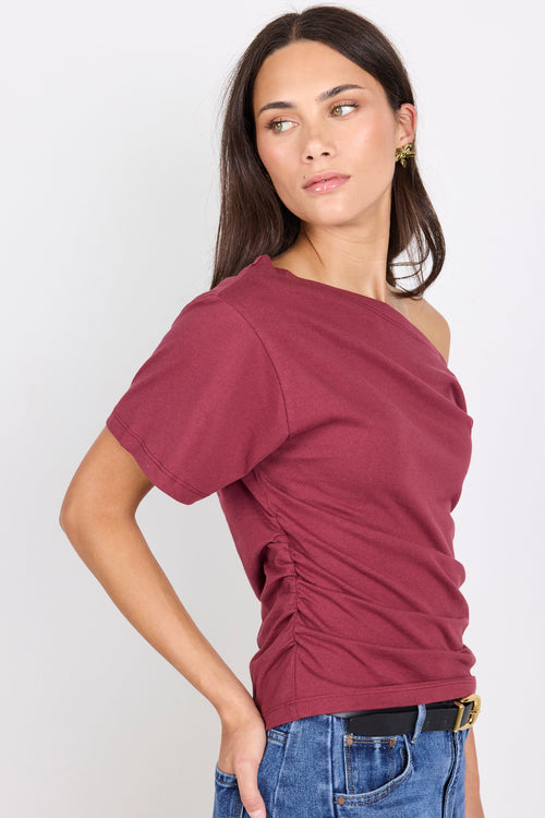 A woman wearing a short-sleeve, off-the-shoulder ruched t-shirt in burgundy, paired with blue jeans and a black belt, photographed against a light gray studio background.
