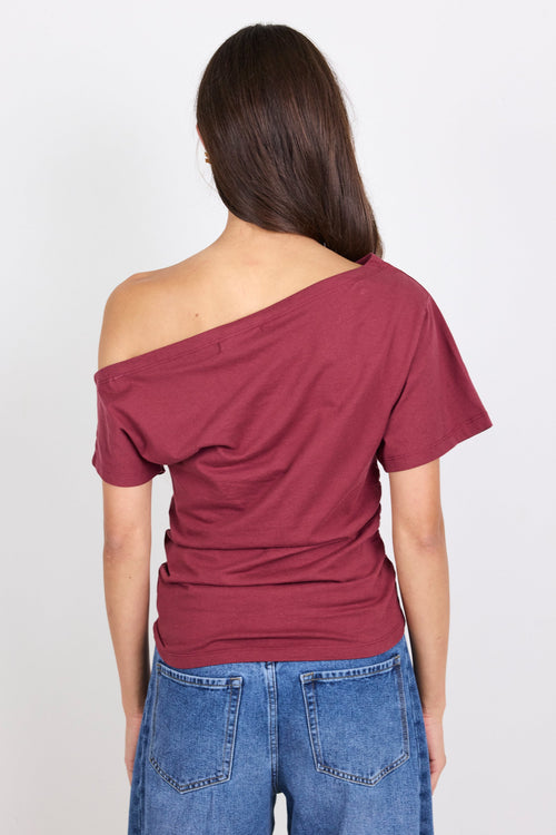 A woman viewed from the back wearing a short-sleeve off-the-shoulder top in burgundy and blue jeans, against a plain white studio background.