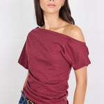 A woman wearing a short-sleeve off-the-shoulder top in burgundy, paired with blue jeans and a black belt, photographed against a light gray studio background.