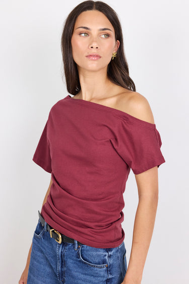 A woman wearing a short-sleeve off-the-shoulder top in burgundy, paired with blue jeans and a black belt, photographed against a light gray studio background.